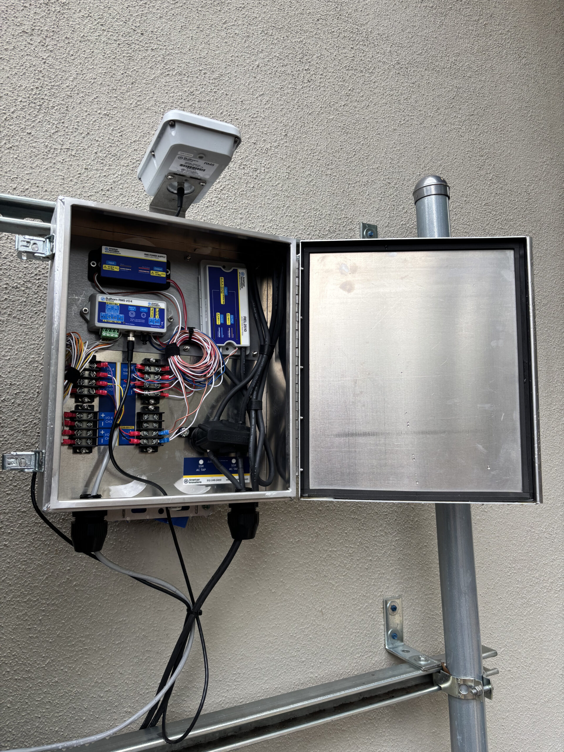 Open stainless steel outdoor enclosure mounted on a wall, showing pre-wired Bullhorn RM510 components including the I/O module, power supply, and REL2510 Solid-State Relay with connected wiring. The enclosure door is open, revealing the internal components. A cellular antenna is mounted on top. The unit is installed on a metal pipe rack against a stucco exterior wall.